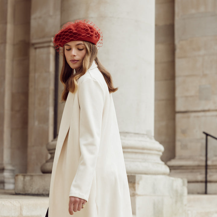 Woman with brown wearing a light coat and a burnt orange wool beret with veiling