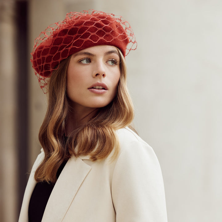 Woman with brown wearing a light coat and a burnt orange wool beret with veiling
