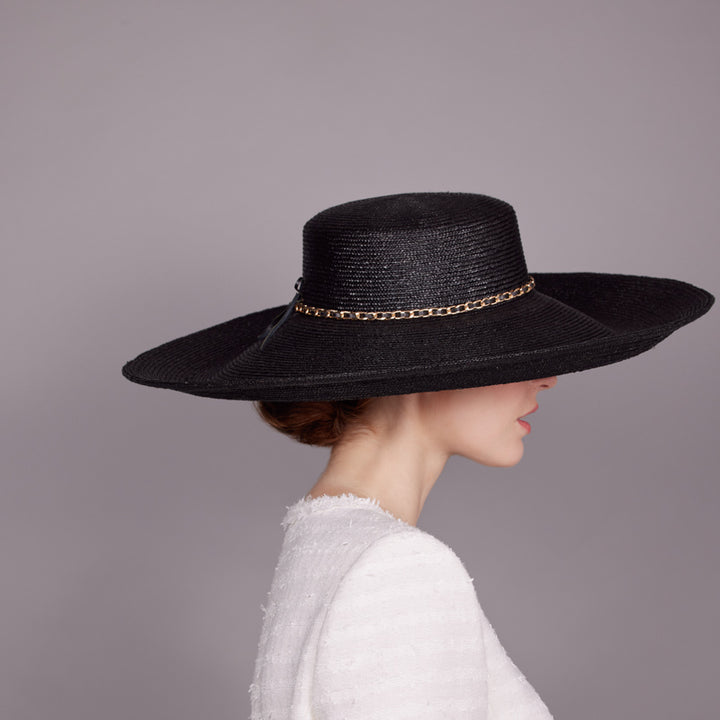 Woman with red hair wearing a white tweed jacket and a black Italian straw hat with a gold chain details