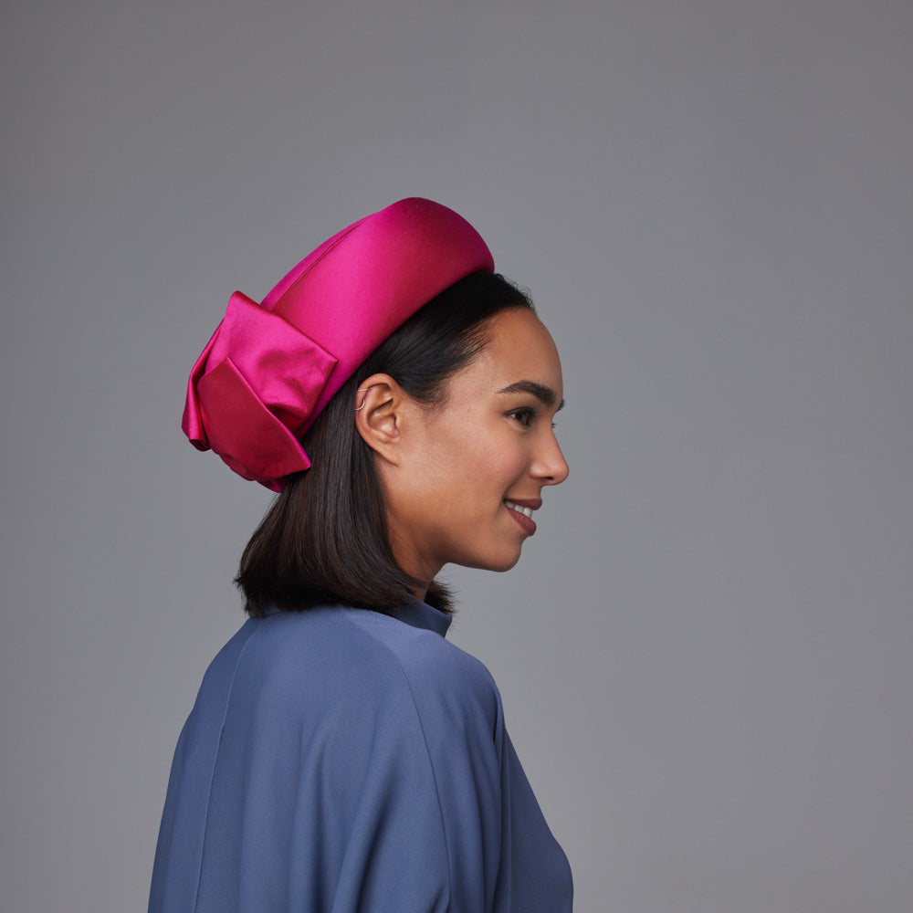 Woman with dark hair wearing a grey shirt and a pink taffeta silk Jackie O pillbox with bows