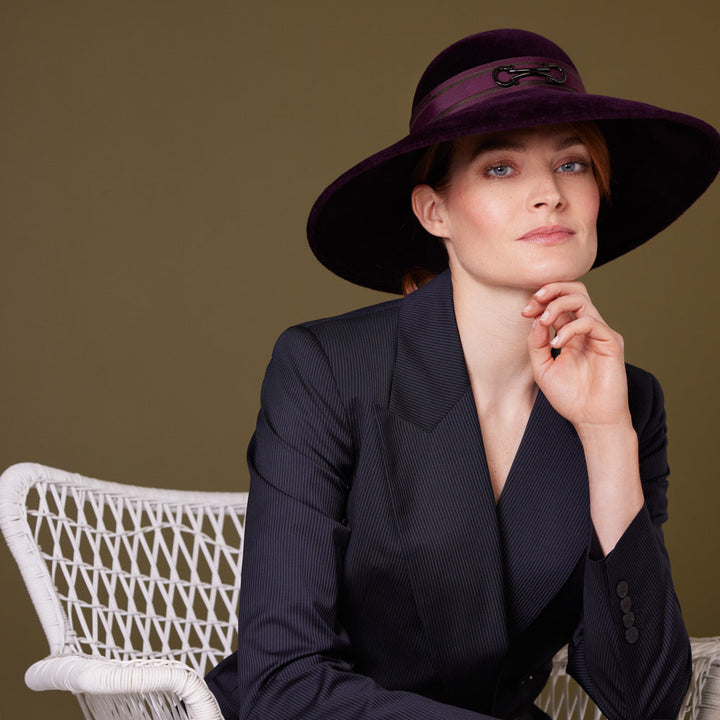 Woman with red hair wearing a navy pin-striped suit and au aubergine velour felt downbrim with a snaffle bit detail on a layered grosgrain band and is sitting on a chair