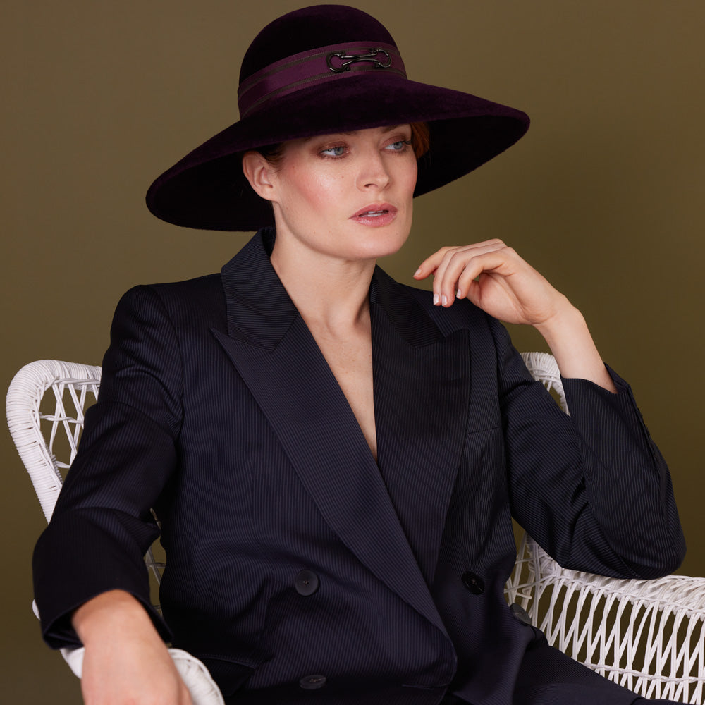Woman with red hair wearing a navy pin-striped suit and au aubergine velour felt downbrim with a snaffle bit detail on a layered grosgrain band and is sitting on a chair