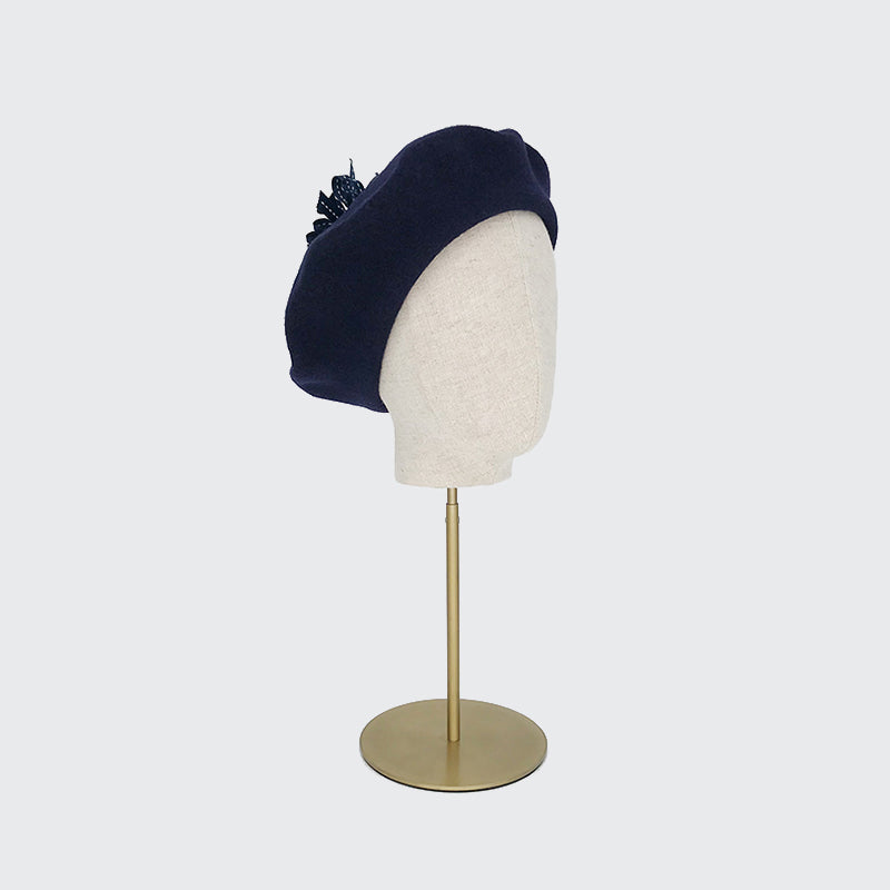 Photo of a navy wool beret with a navy ribbon pom on a linen display head