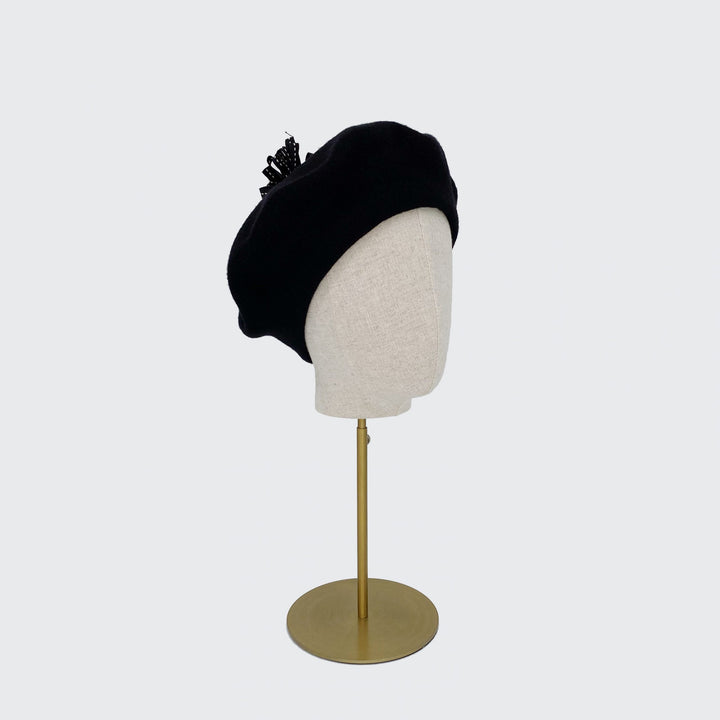 Photo of a black wool beret with a black ribbon pom on a linen display head