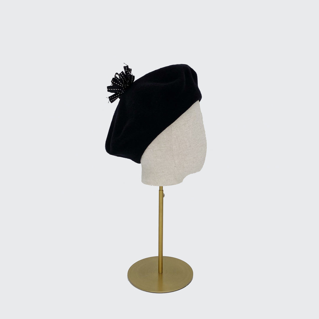 Side view of a black wool beret with a black ribbon pom on a linen display head