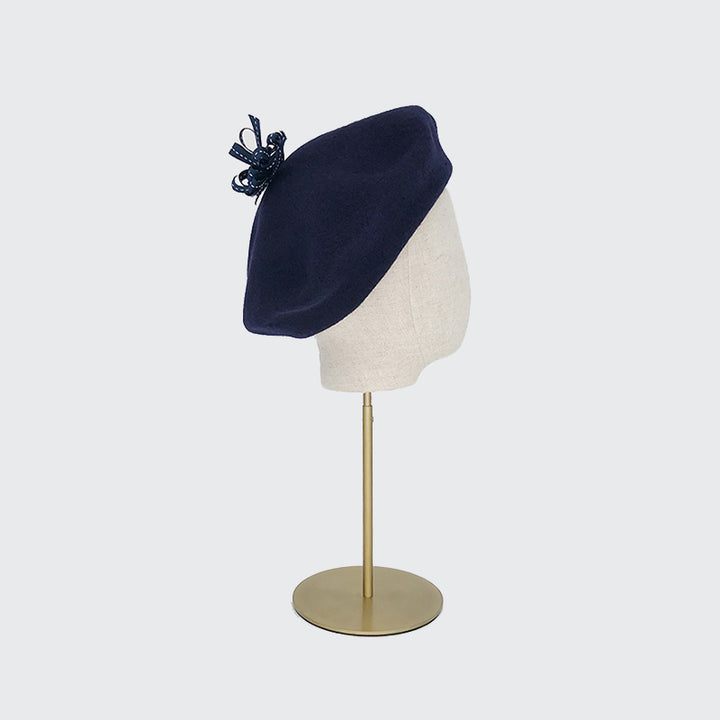 Side view of a navy wool beret with a navy ribbon pom on a linen display ehad
