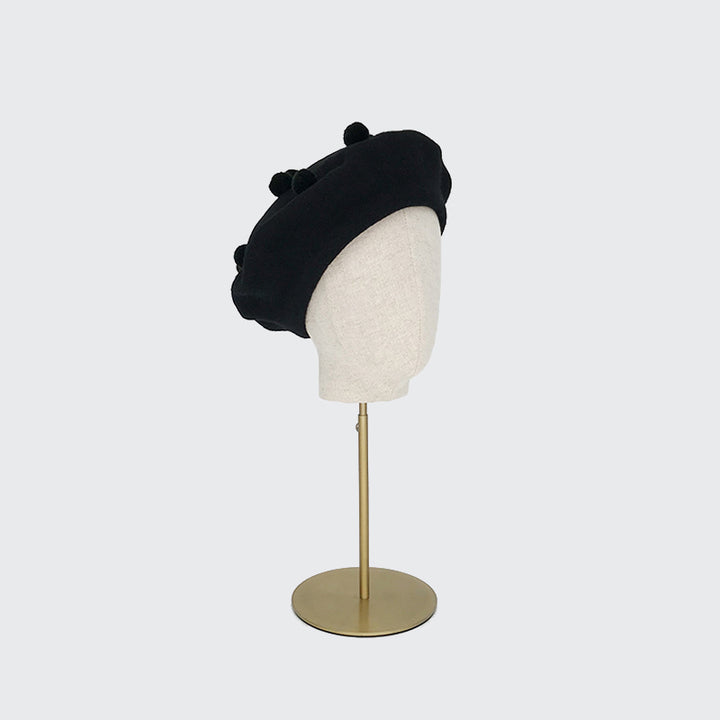 Photo of a black wool beret with black pom poms on a linen display head