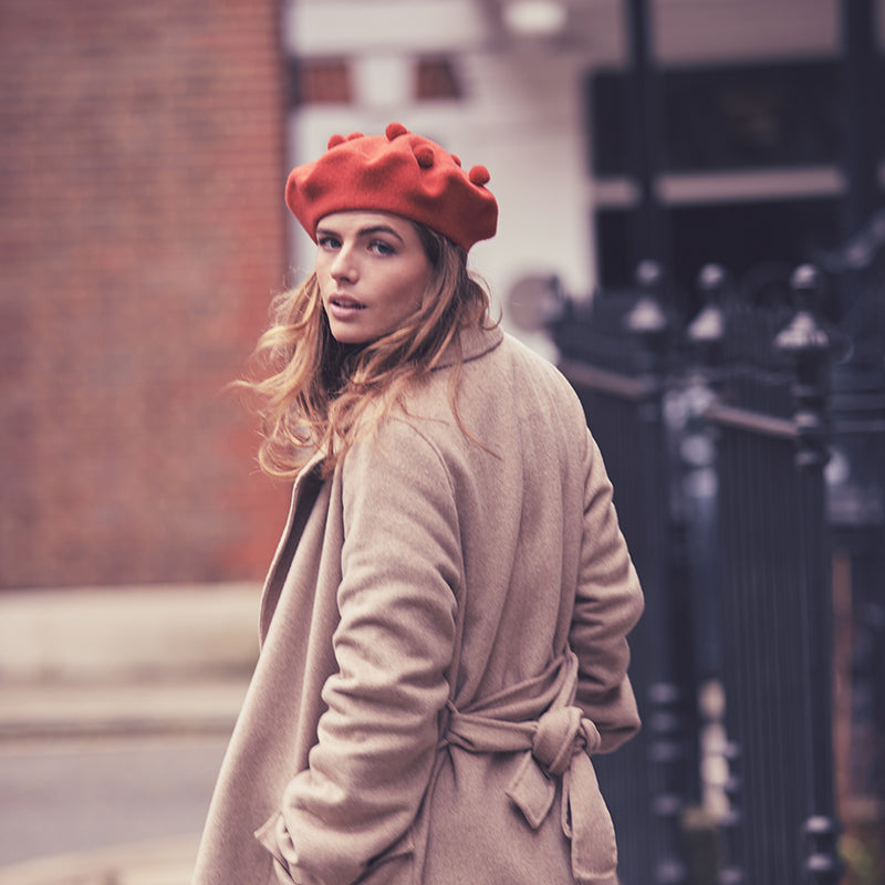 Woman with long hair wearing a camel coat and a burnt orange pom pom beret