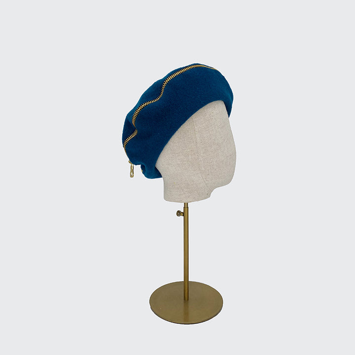 Side view of a teal wool beret with gold zip on a linen display head