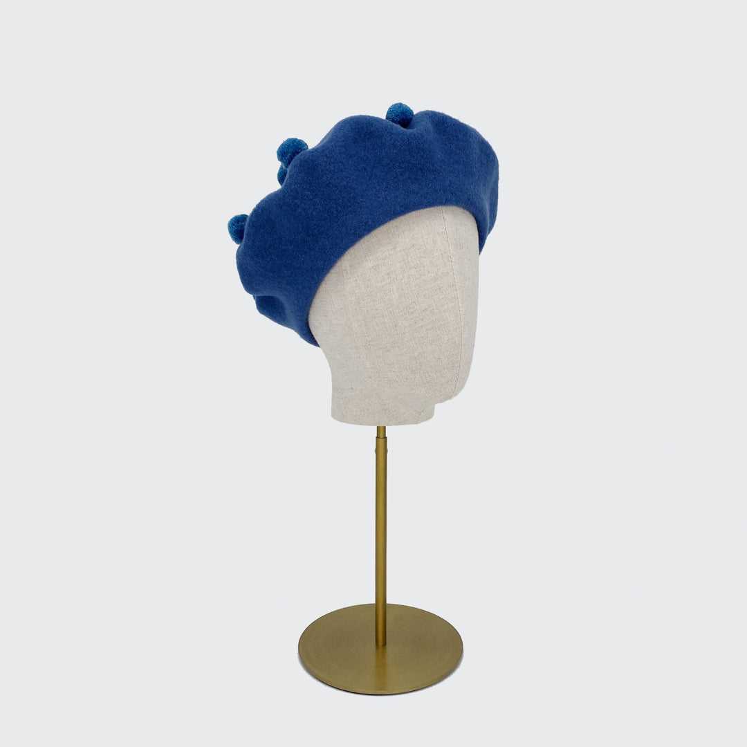 Photo of a denim blue wool beret with denim blue pom poms on a linen display head