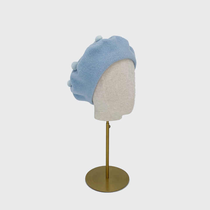 Photo of a pale blue wool beret with pale blue pom poms on a linen display head