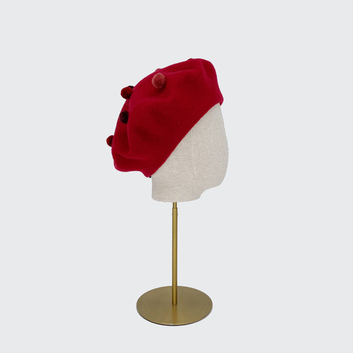 Photo of a dark red wool beret with red pom poms on a linen display head