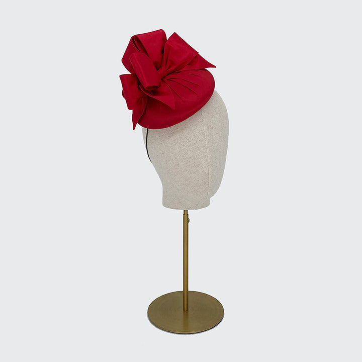 Side view of a red silk pillbox with bows on a linen display head