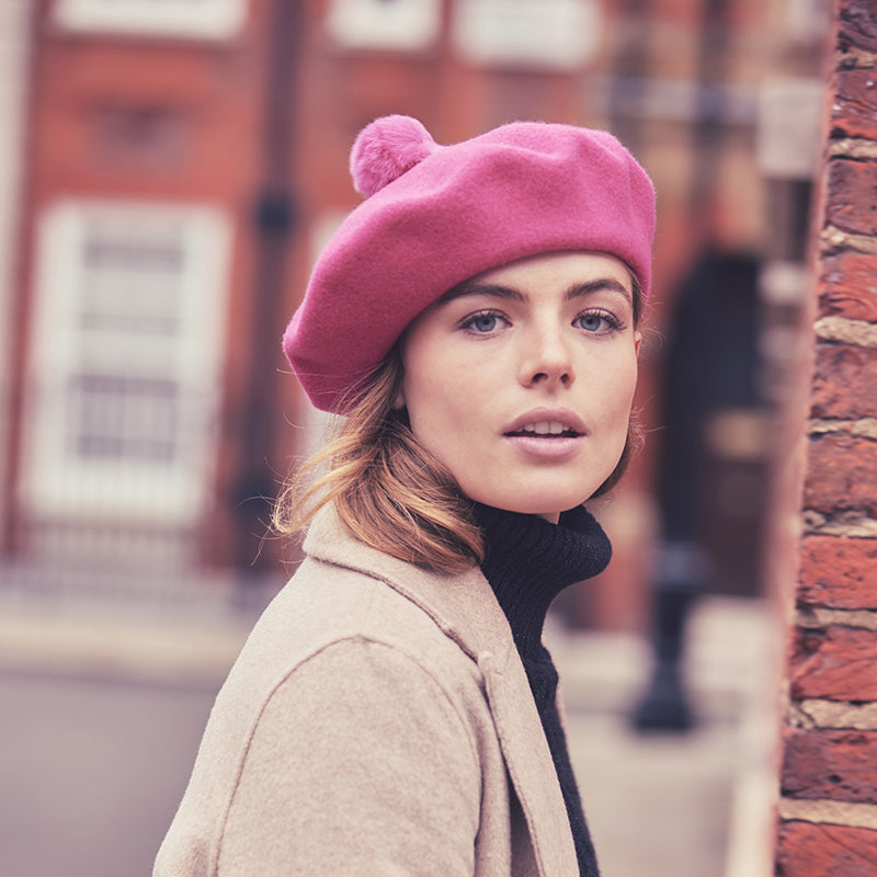 Woman with long hair wearing a camel coat and a hot pink beret with a fur pom