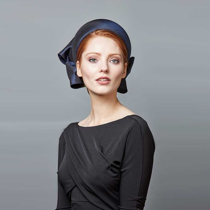 Woman with red hair wearing a black dress and a navy taffeta silk Jackie O pillbox with bows