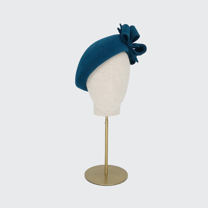 Side view of a teal velour felt beret with bows on a linen display head