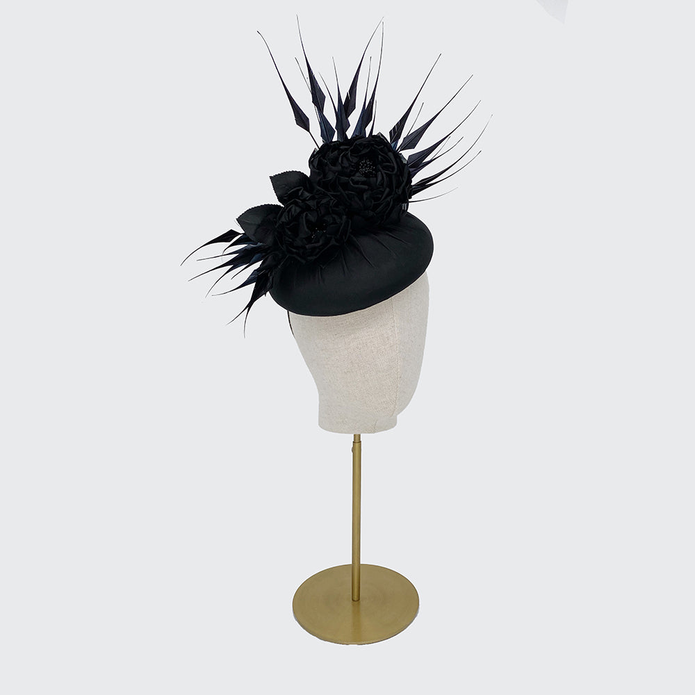 Side view of a black large silk pillbox with silk roses and diamond-cut feathers on a linen display head