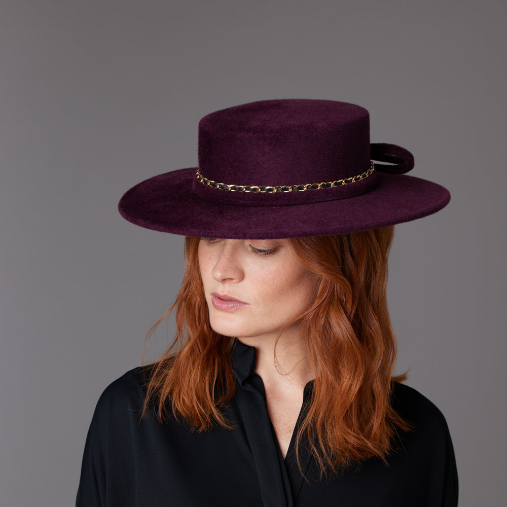 Woman with red hair wearing a black shirt and an aubergine velour felt boater with a gold chain detail