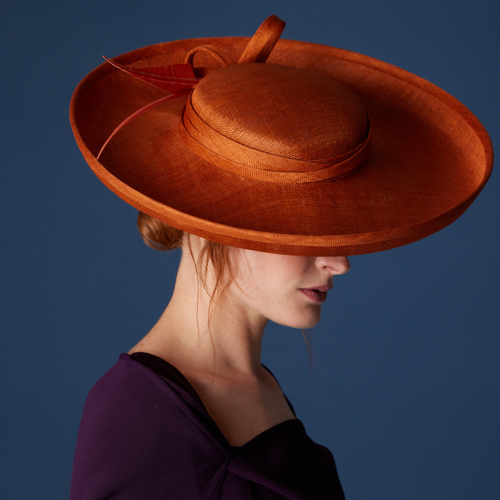 Woman with red hair wearing purple dress and a burnt orange Breton hat with sharp feathers