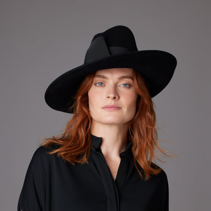 Woman with red hair wearing a black blouse and a black high crown velour felt fedora with wide grosgrain band