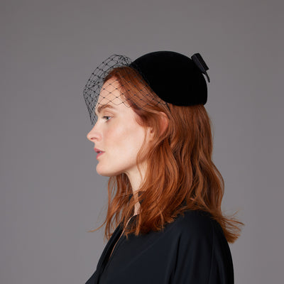 Woman with red hair wearing a black shirt and a black velvet caplet with a face veil and a bow