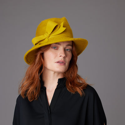 Woman with red hair wearing a black blouse and a mustard melusine trilby with bow