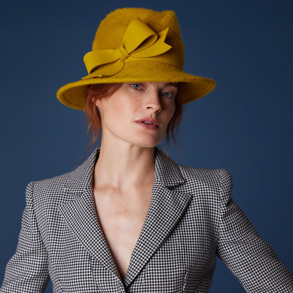 Woman with red hair wearing a houndstooth blazer and a mustard melusine trilby with bow