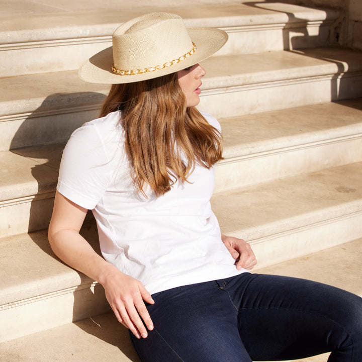 Woman with long hair wearing jeans and a white tshirt and a wide brim fedora with a gold chain detail