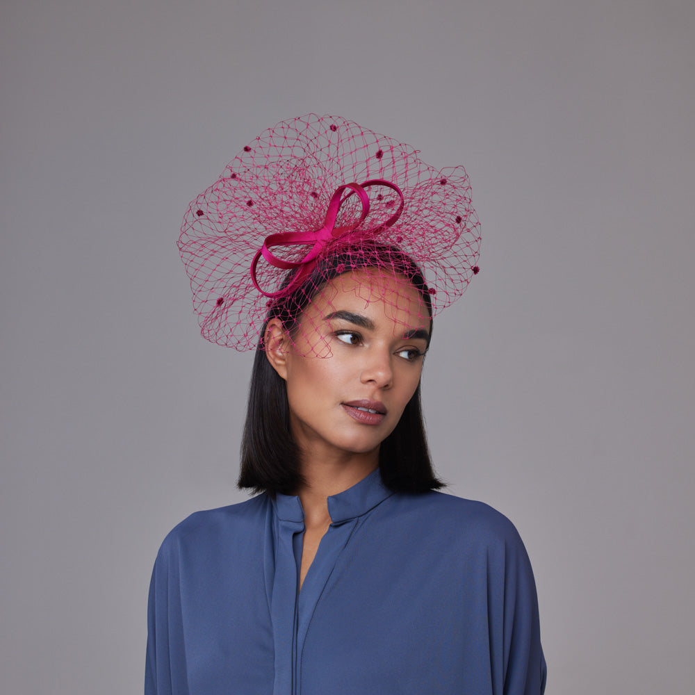 Woman with darj hair wearing a blue shirt and a deep pink looped bow headband with spotty veiling